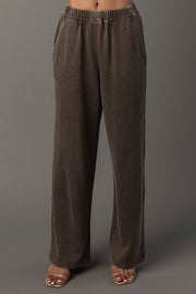 A person wearing Panache Apparel Collection’s Cocoa Washed Soft Sweatpants, featuring a loose fit, wide legs, side pockets, and an elastic waistband, paired with open-toe heels, stands against a plain gray background.