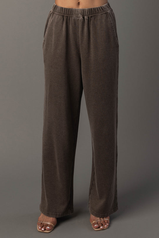 A person wearing Panache Apparel Collection’s Cocoa Washed Soft Sweatpants, featuring a loose fit, wide legs, side pockets, and an elastic waistband, paired with open-toe heels, stands against a plain gray background.