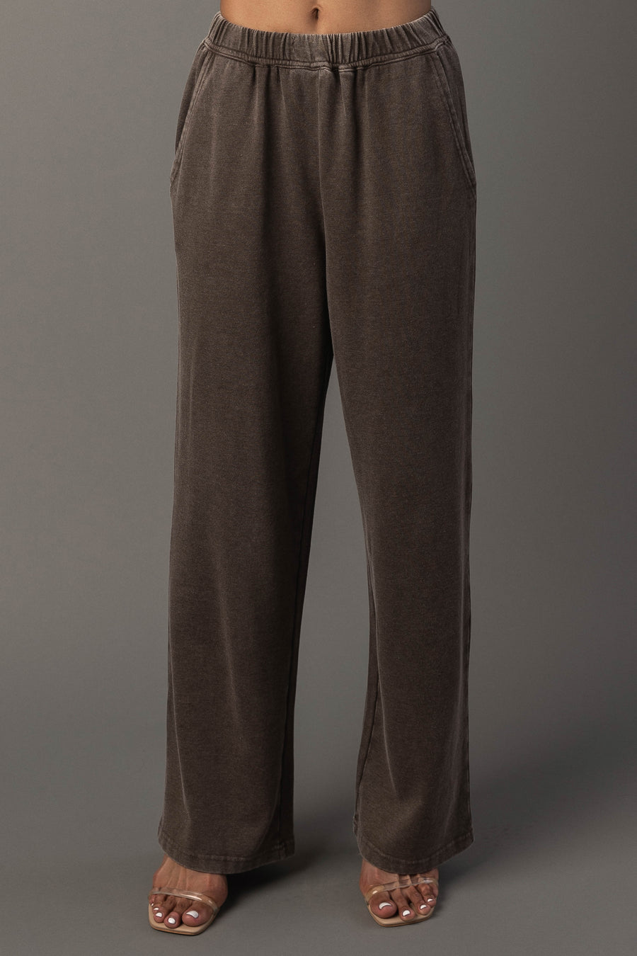 A person wearing Panache Apparel Collection’s Cocoa Washed Soft Sweatpants, featuring a loose fit, wide legs, side pockets, and an elastic waistband, paired with open-toe heels, stands against a plain gray background.