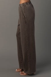 A person models the Panache Apparel Collection’s Cocoa Washed Soft Sweatpants. A high-waisted, loose-fitting dark brown style featuring an elastic waistband and convenient side pockets, paired with heeled sandals and posed sideways against a plain gray background.