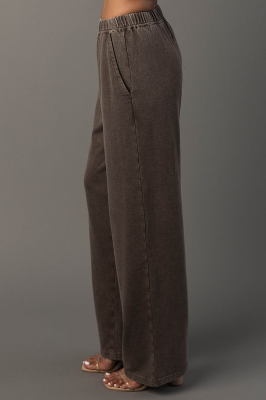A person models the Panache Apparel Collection’s Cocoa Washed Soft Sweatpants. A high-waisted, loose-fitting dark brown style featuring an elastic waistband and convenient side pockets, paired with heeled sandals and posed sideways against a plain gray background.
