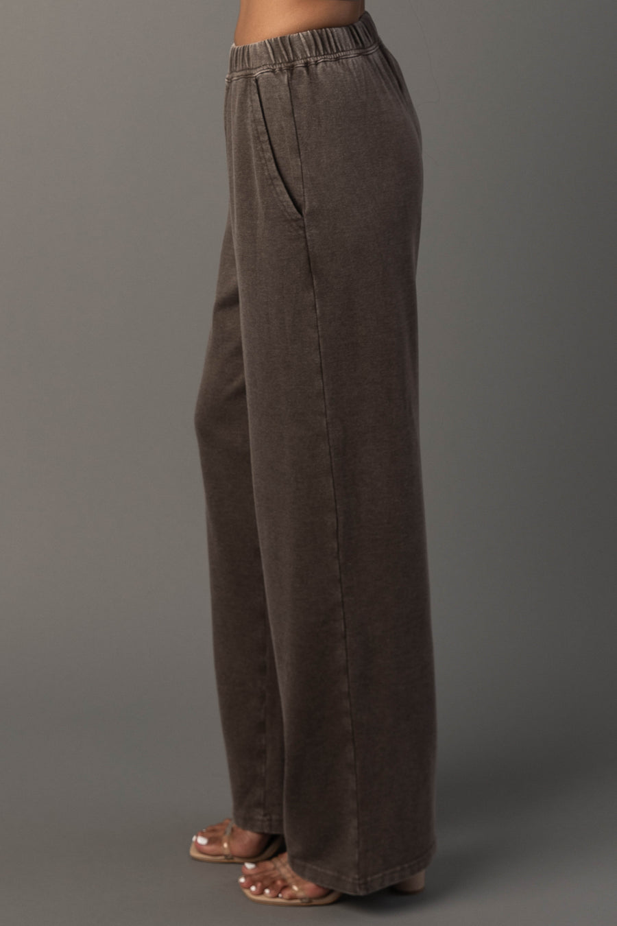 A person models the Panache Apparel Collection’s Cocoa Washed Soft Sweatpants. A high-waisted, loose-fitting dark brown style featuring an elastic waistband and convenient side pockets, paired with heeled sandals and posed sideways against a plain gray background.