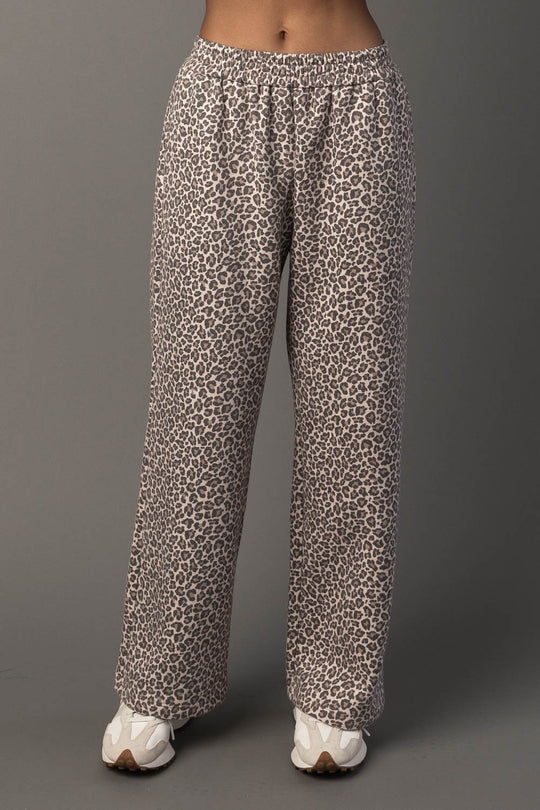 A person wearing Panache Apparel Collection’s Cream & Brown Leopard Sweatpants and white sneakers stands against a plain gray background. The photo shows the sweatpants from waist to feet.