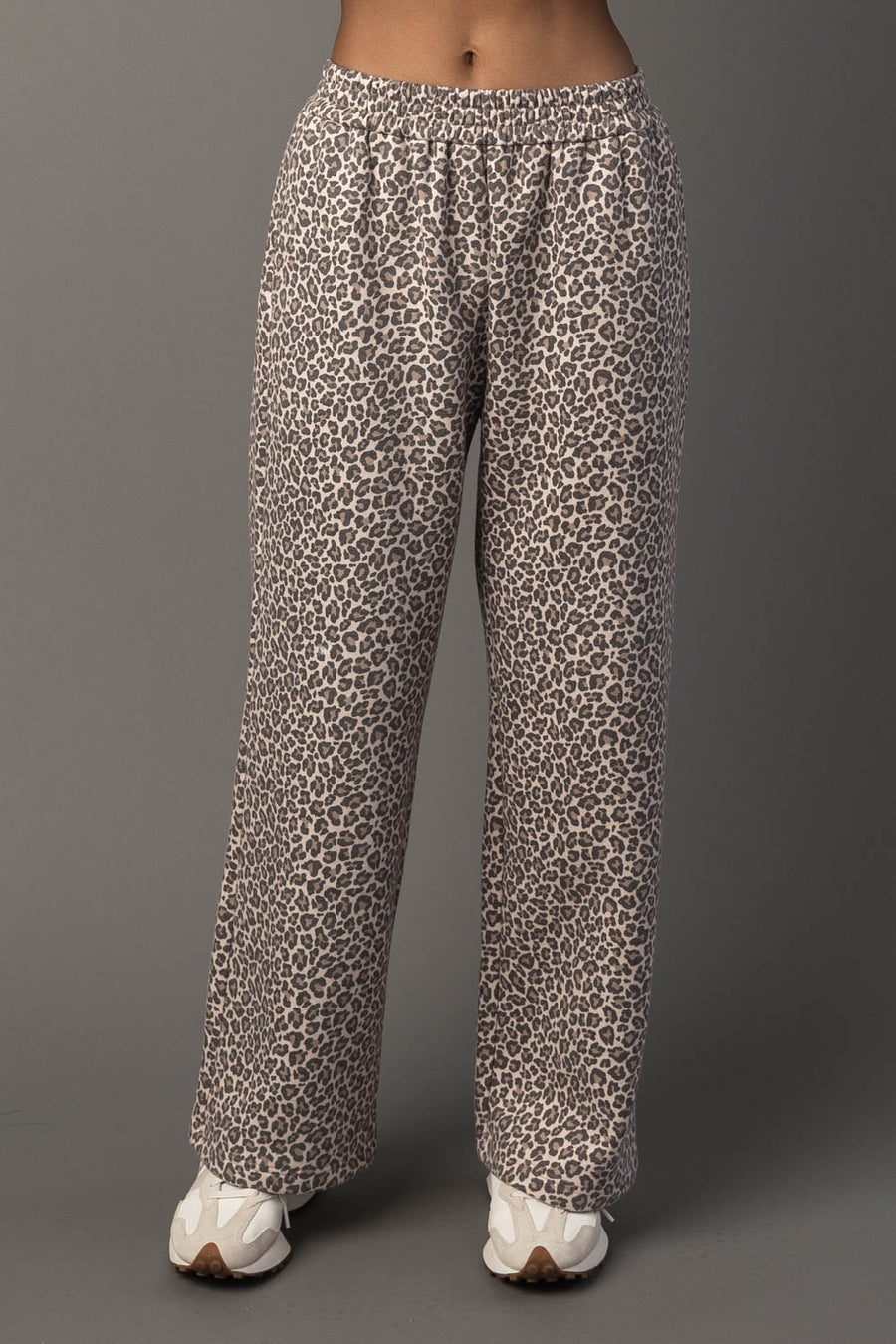 A person wearing Panache Apparel Collection’s Cream & Brown Leopard Sweatpants and white sneakers stands against a plain gray background. The photo shows the sweatpants from waist to feet.