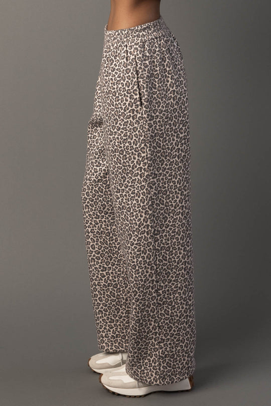 A person stands sideways against a plain gray background wearing Panache Apparel Collection’s Cream & Brown Leopard Sweatpants and white shoes.