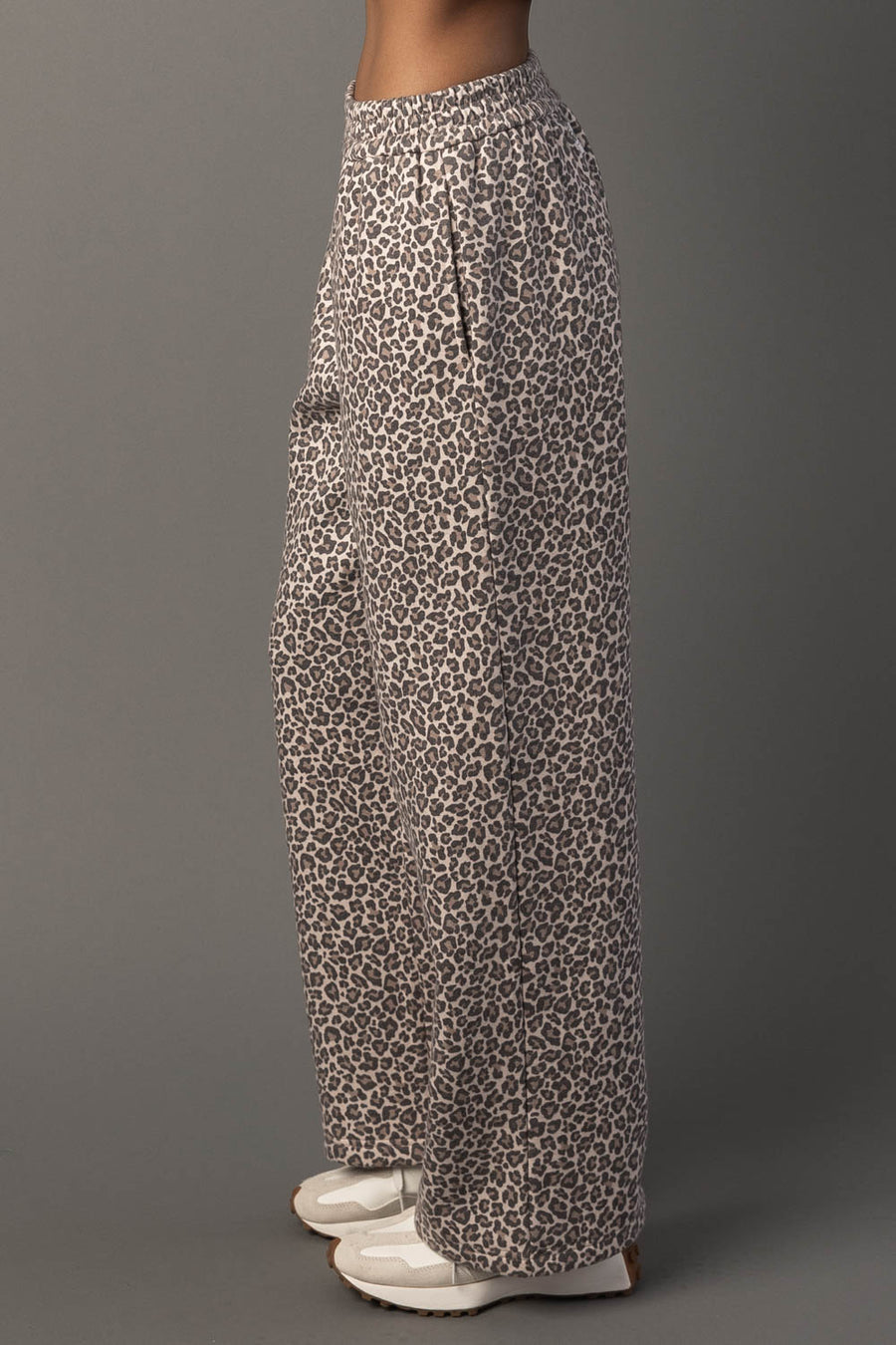 A person stands sideways against a plain gray background wearing Panache Apparel Collection’s Cream & Brown Leopard Sweatpants and white shoes.
