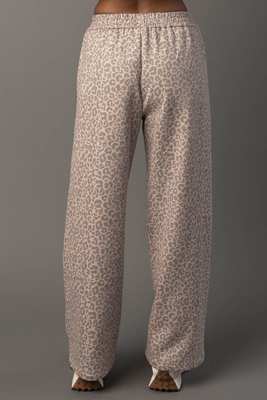 A person wearing Panache Apparel Collection’s Blush & Light Brown Leopard Sweatpants, featuring a loose fit and high-waisted design with a blush and light brown leopard print, stands facing away against a plain grey background.