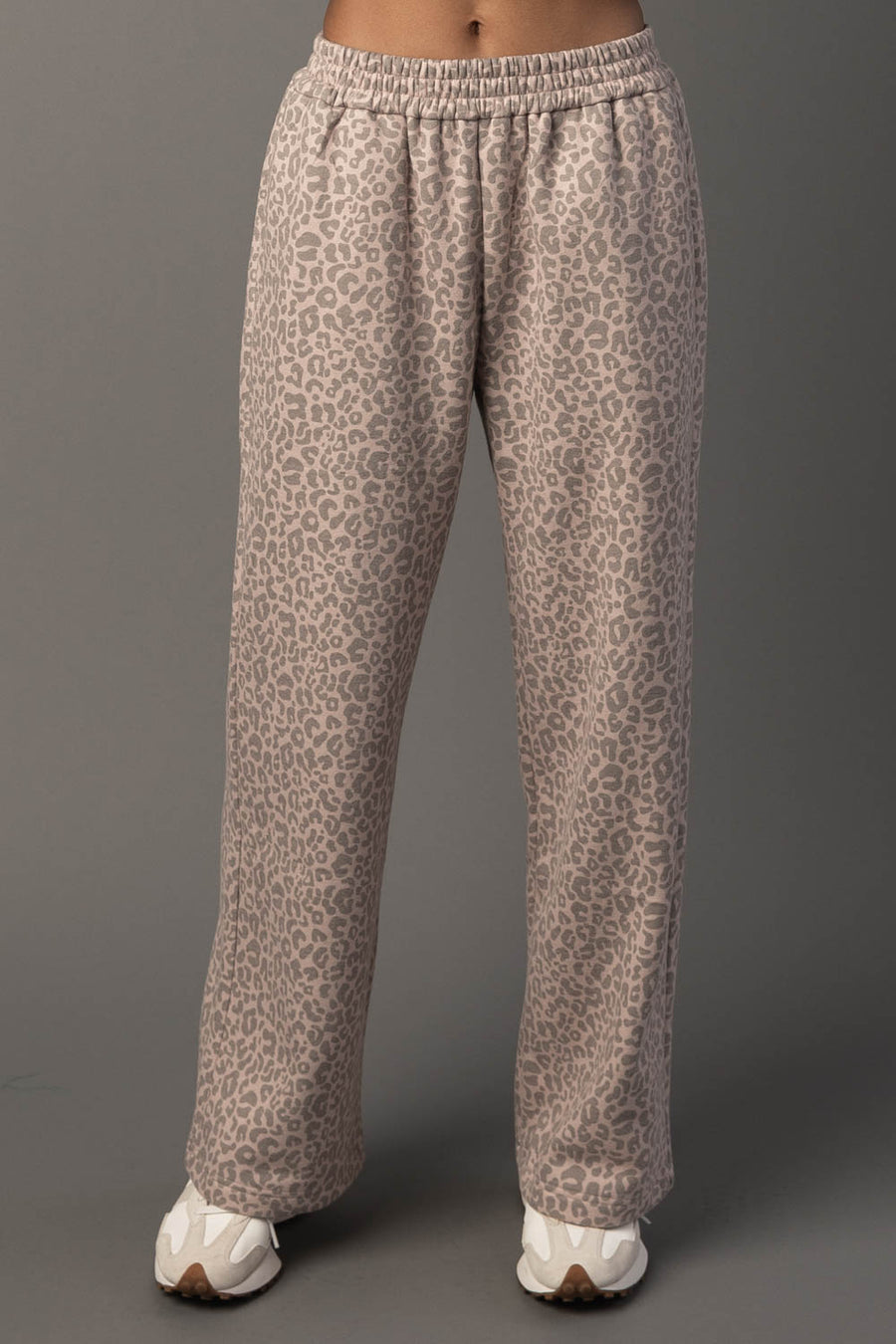 A person is wearing Blush & Light Brown Leopard Sweatpants by Panache Apparel Collection, paired with white sneakers, and standing against a plain gray background. The photo captures the sweatpants from waist to feet.