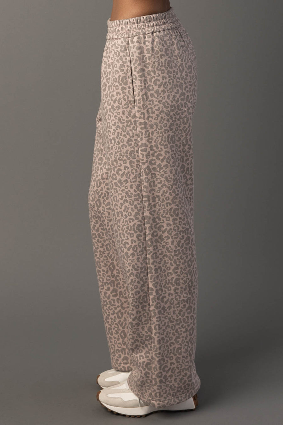 A person is wearing the Panache Apparel Collection’s Blush & Light Brown Leopard Sweatpants, featuring a loose, high-waisted fit with an elastic waistband and a gray leopard print pattern. They are styled with white platform shoes against a gray background, with the side view highlighting the unique fit of the pants.
