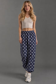 A woman with long blond hair and a neutral expression stands against a gray background, modeling the Panache Apparel Collection Navy & White Polka Dot Sweatpants. She pairs them with a beige sleeveless crop top and white sneakers.