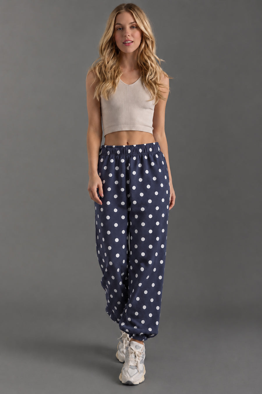 A woman with long blond hair and a neutral expression stands against a gray background, modeling the Panache Apparel Collection Navy & White Polka Dot Sweatpants. She pairs them with a beige sleeveless crop top and white sneakers.