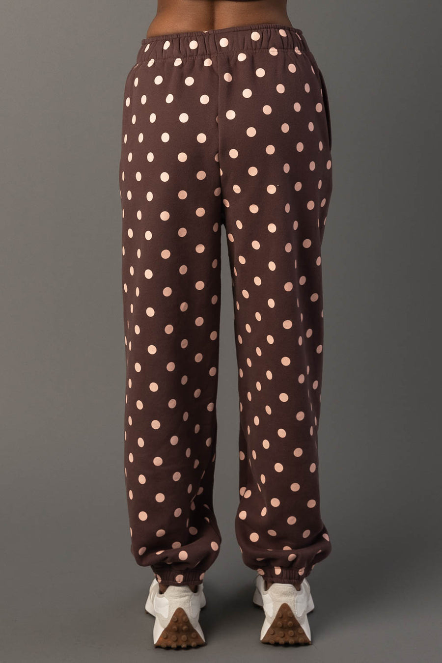 A person is seen from the back wearing Panache Apparel Collection’s Brown & Light Pink Polka Dot Sweatpants, featuring an elastic waistband and cuffs. They are paired with white sneakers and photographed against a plain gray background.