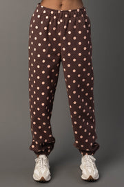 A person wearing Panache Apparel Collection’s Brown & Light Pink Polka Dot Sweatpants and white sneakers stands against a plain grey background.
