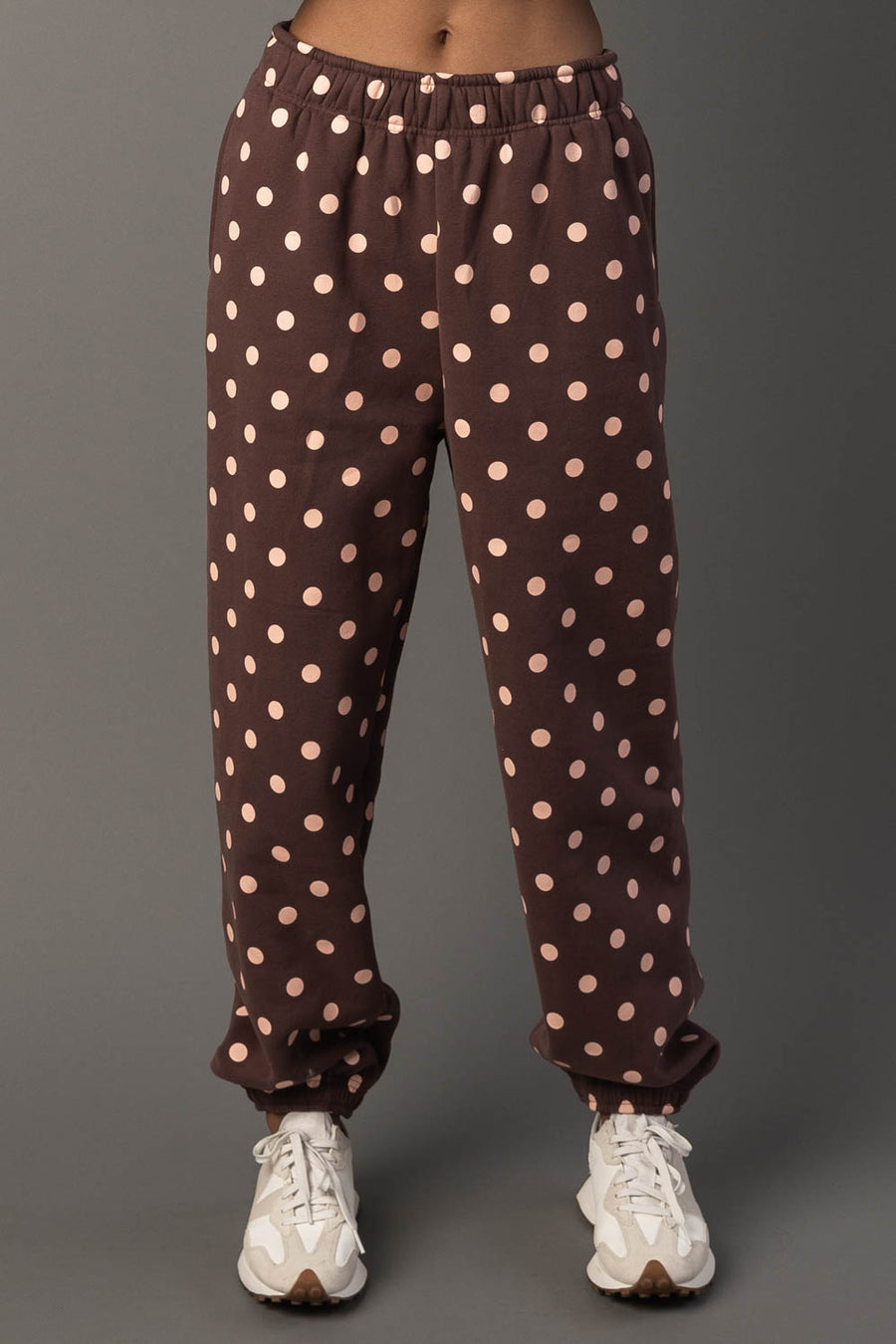 A person wearing Panache Apparel Collection’s Brown & Light Pink Polka Dot Sweatpants and white sneakers stands against a plain grey background.
