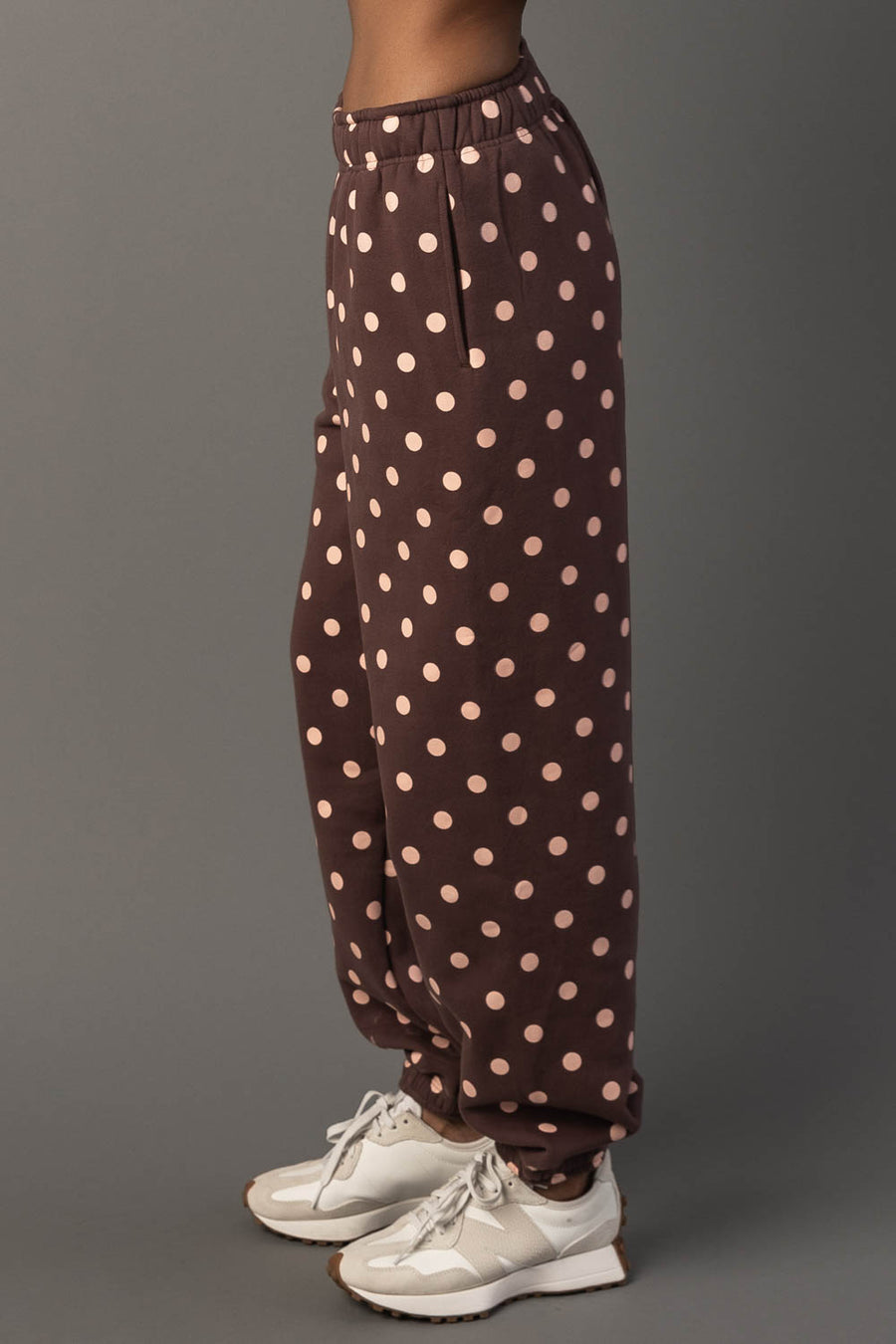 A person models the Panache Apparel Collection Brown & Light Pink Polka Dot Sweatpants with white sneakers, standing sideways against a gray background.