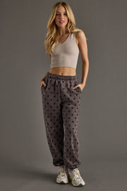 A woman with long blonde hair stands with her hands in her pockets against a plain gray background, wearing a beige crop tank top, white sneakers, and the Panache Apparel Collection’s Dark Gray & Black Polka Dot Sweatpants. Draped casually over her arm is an oversized fleece hoodie, adding a cozy touch to her relaxed look.