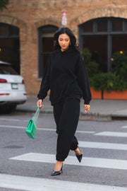 A person in a black hooded sweatshirt and Black Panache Sweatpants walks across a crosswalk, carrying a bright green quilted handbag and wearing black slingback heels.