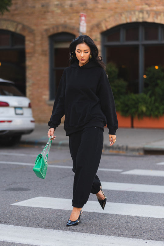 A person in a black hooded sweatshirt and Black Panache Sweatpants walks across a crosswalk, carrying a bright green quilted handbag and wearing black slingback heels.