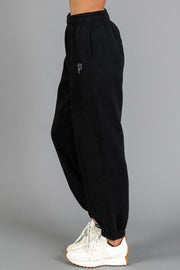A person wearing Black Panache Sweatpants from Panache Apparel Collection and white sneakers stands sideways against a plain gray background.