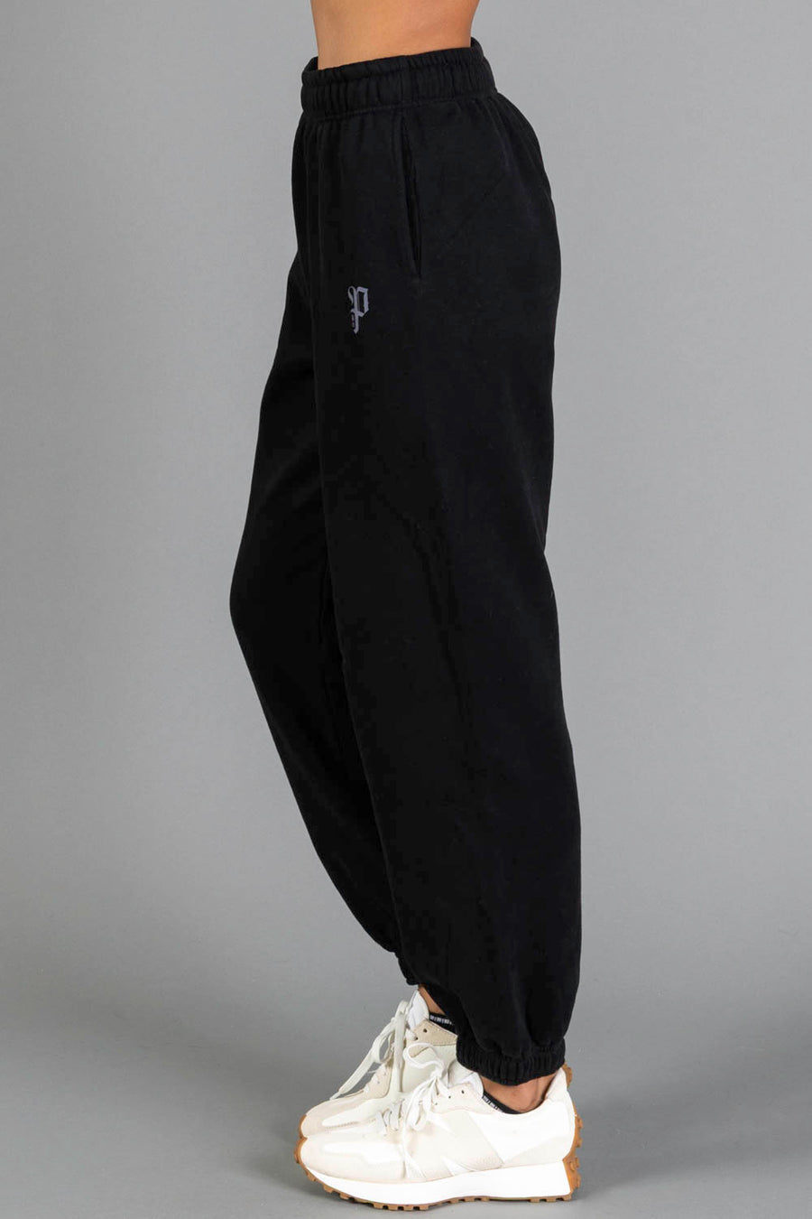 A person wearing Black Panache Sweatpants from Panache Apparel Collection and white sneakers stands sideways against a plain gray background.