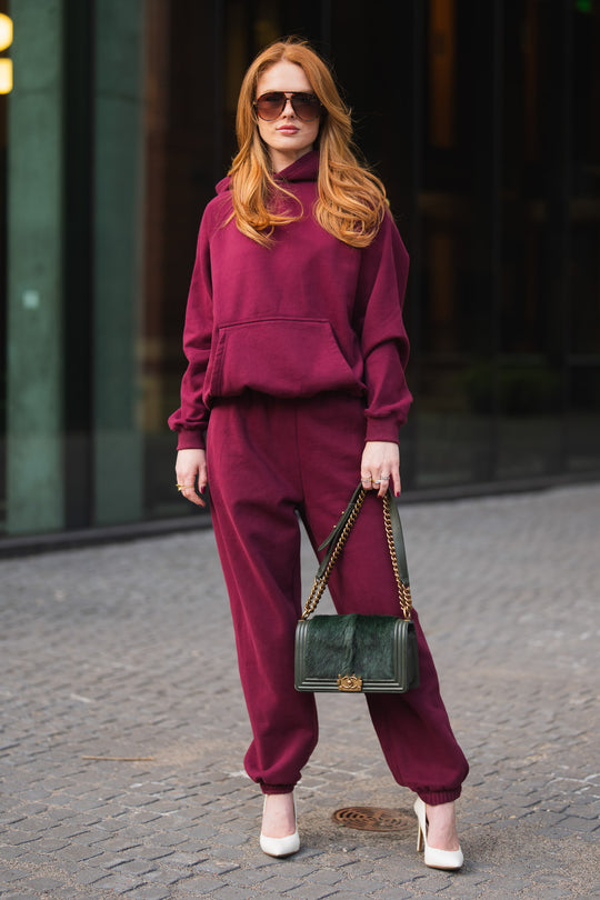 A woman wearing a matching maroon hooded sweatshirt and Maroon Panache Sweatpants, white high heels, and holding a dark green shoulder bag.