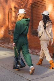 Two women wearing matching hooded sweatshirts and Dark Green Panache Sweatpants walk along a brick wall, each wearing a baseball cap.