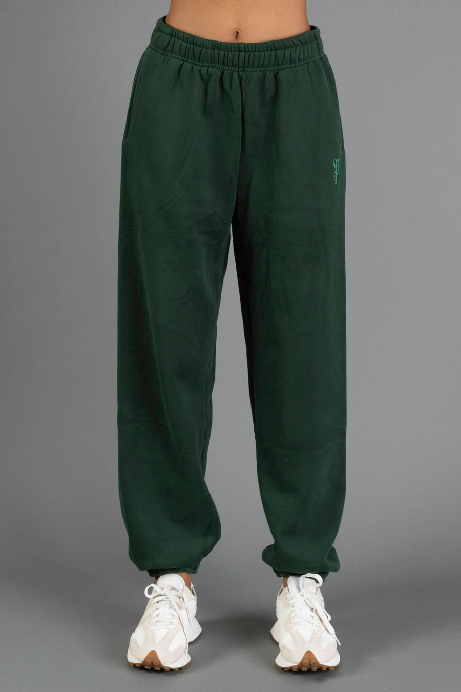 A person is wearing the Panache Apparel Collection’s Dark Green Panache Sweatpants, featuring an elastic waistband and cuffs, paired with white sneakers and standing against a plain gray background.