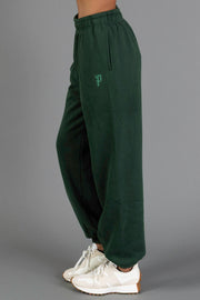 A person stands sideways wearing the Dark Green Panache Sweatpants from the Panache Apparel Collection, featuring an embroidered logo, paired with white sneakers, against a plain gray background.