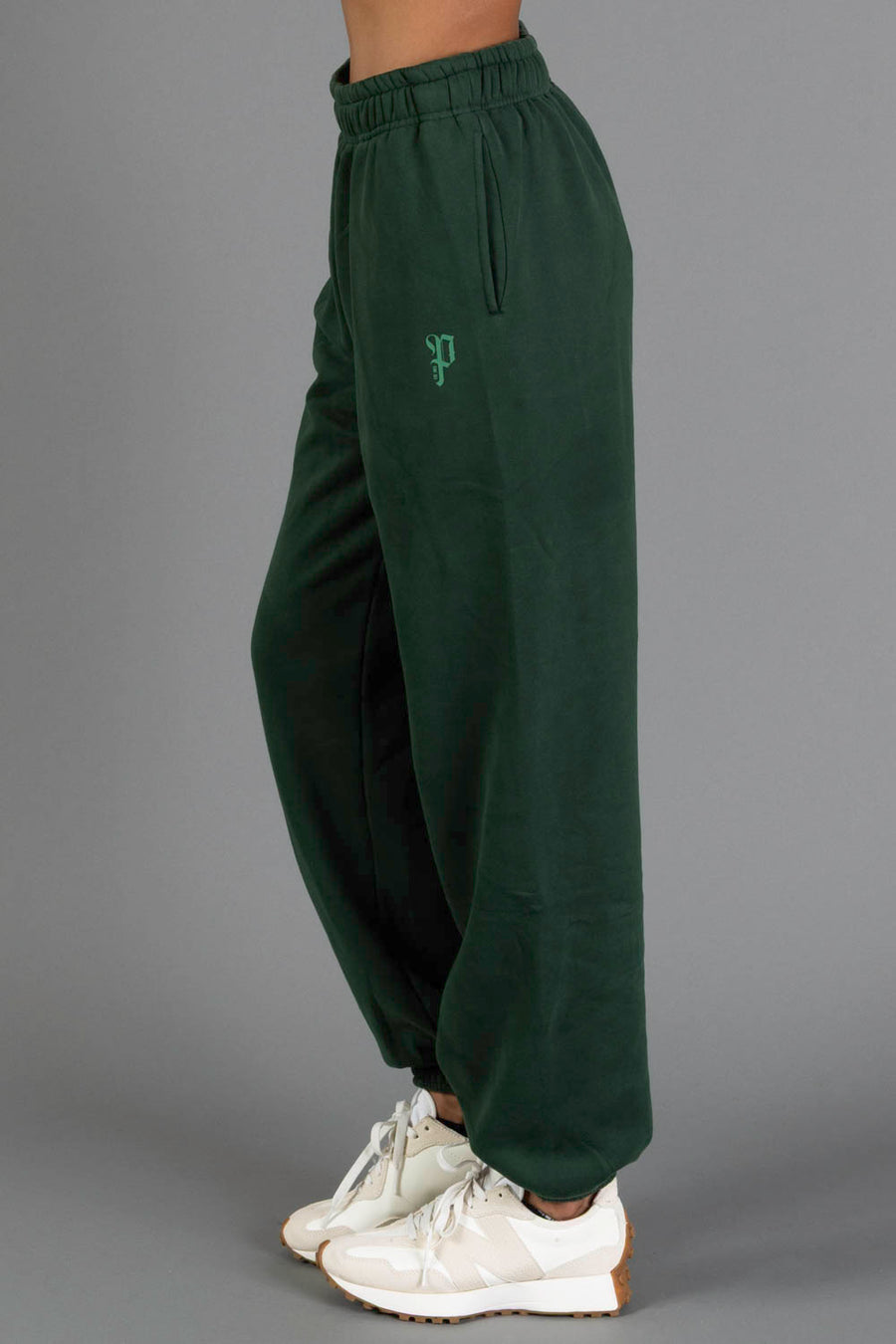 A person stands sideways wearing the Dark Green Panache Sweatpants from the Panache Apparel Collection, featuring an embroidered logo, paired with white sneakers, against a plain gray background.