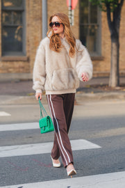 A woman wearing a fuzzy beige hooded top and Brown Lounge Sweatpants from Panache Apparel Collection with side stripes walks across a city crosswalk while carrying a small green handbag.