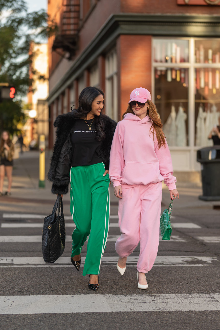 Two women walk across a city crosswalk; one wears a black top and Green Lounge Sweatpants by Panache Apparel Collection, and the other wears a matching pink hoodie and sweatpants with a pink baseball cap.