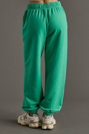 A person, back to the camera, wears Panache Apparel Collection’s bright green Green Panache Sweatpants with an elastic waistband and white sneakers, all set against a plain gray background.