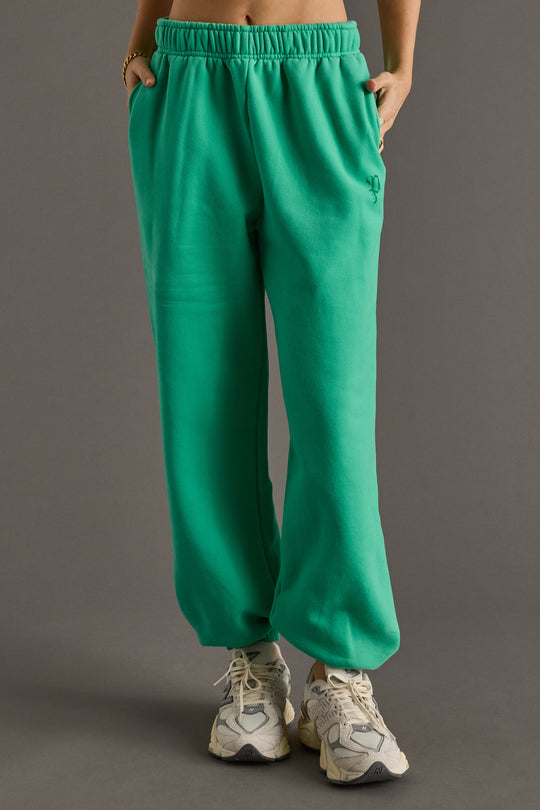 A person models the Green Panache Sweatpants from Panache Apparel Collection, featuring an elastic waistband, paired with white sneakers and hands in pockets. Only the lower body is visible against a plain gray background.
