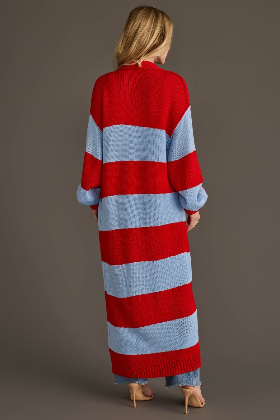 A woman with long blonde hair stands with her back to the camera, wearing the Panache Apparel Collection Red & Light Blue Striped Duster Cardigan, styled with light blue pants and nude heels.