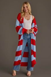A smiling woman from Panache Apparel Collection stands against a plain brown background wearing the Red & Light Blue Striped Duster Cardigan over a white tank top, blue jeans, and heeled sandals.