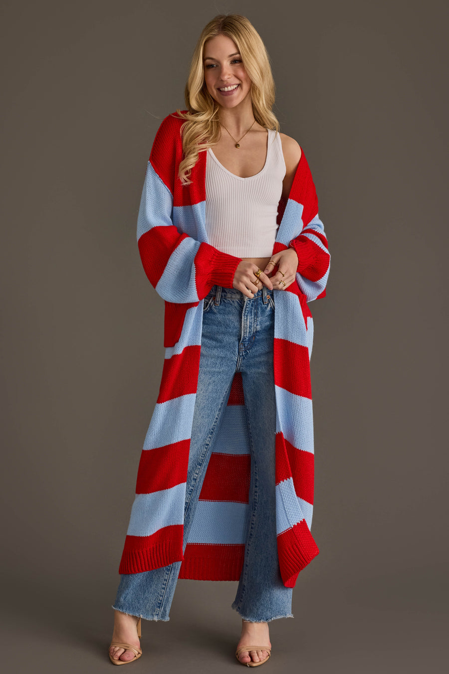 A smiling woman from Panache Apparel Collection stands against a plain brown background wearing the Red & Light Blue Striped Duster Cardigan over a white tank top, blue jeans, and heeled sandals.