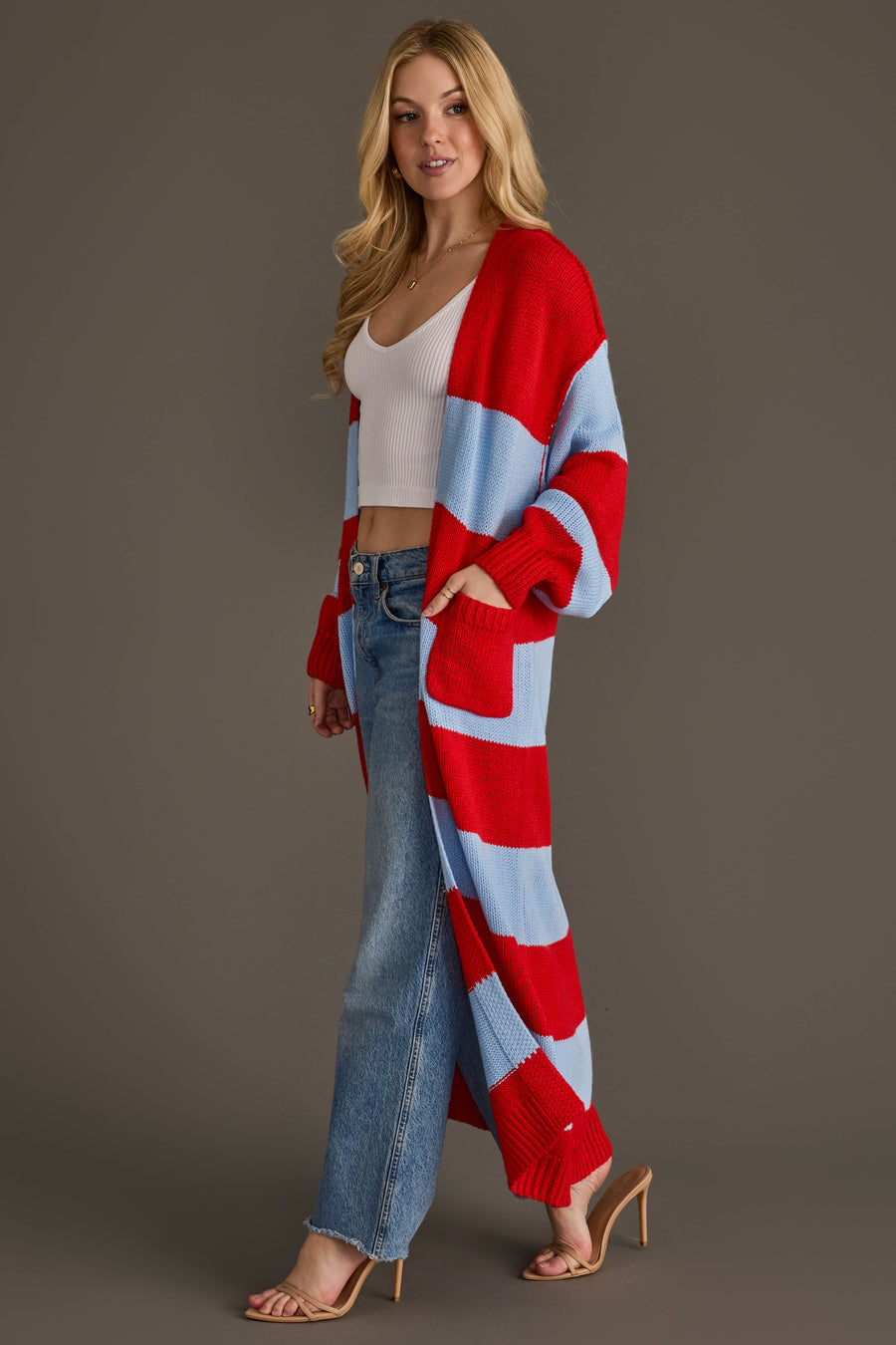 A woman stands against a plain brown background wearing the Panache Apparel Collection Red & Light Blue Striped Duster Cardigan, paired with a white ribbed crop top, blue jeans, and nude heeled sandals.