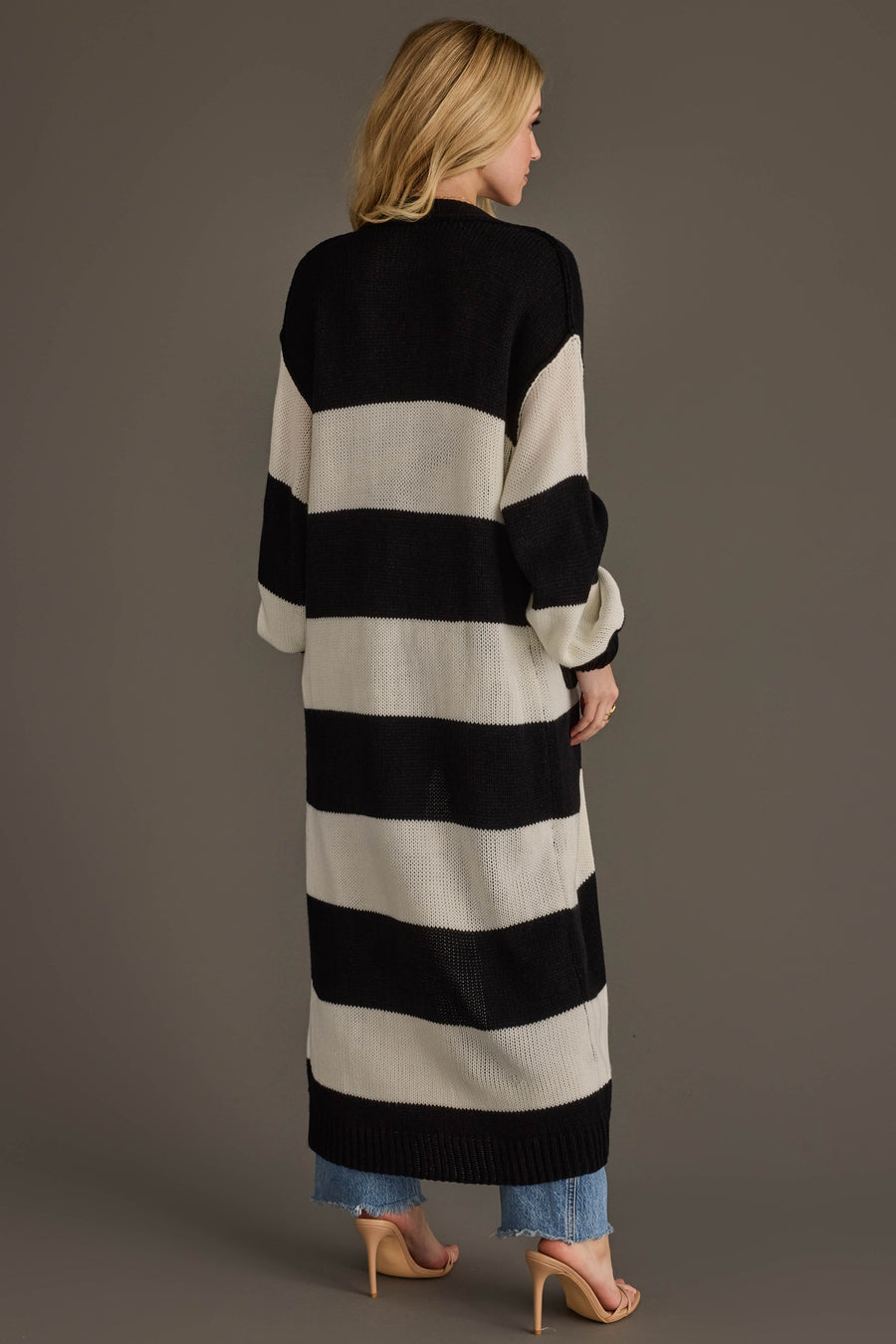 A woman with blonde hair stands with her back to the camera against a plain background, wearing jeans, heels, and the Panache Apparel Collection Black & White Striped Duster Cardigan.