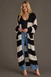 A woman models the Panache Apparel Collection Black & White Striped Duster Cardigan, layering it over a black top and blue jeans as she stands against a plain background with her hands clasped and a slight smile on her face.