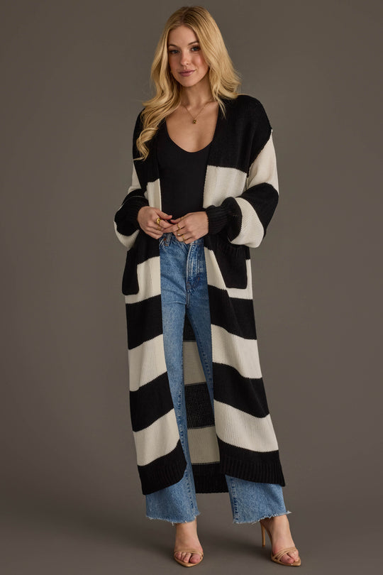 A woman models the Panache Apparel Collection Black & White Striped Duster Cardigan, layering it over a black top and blue jeans as she stands against a plain background with her hands clasped and a slight smile on her face.