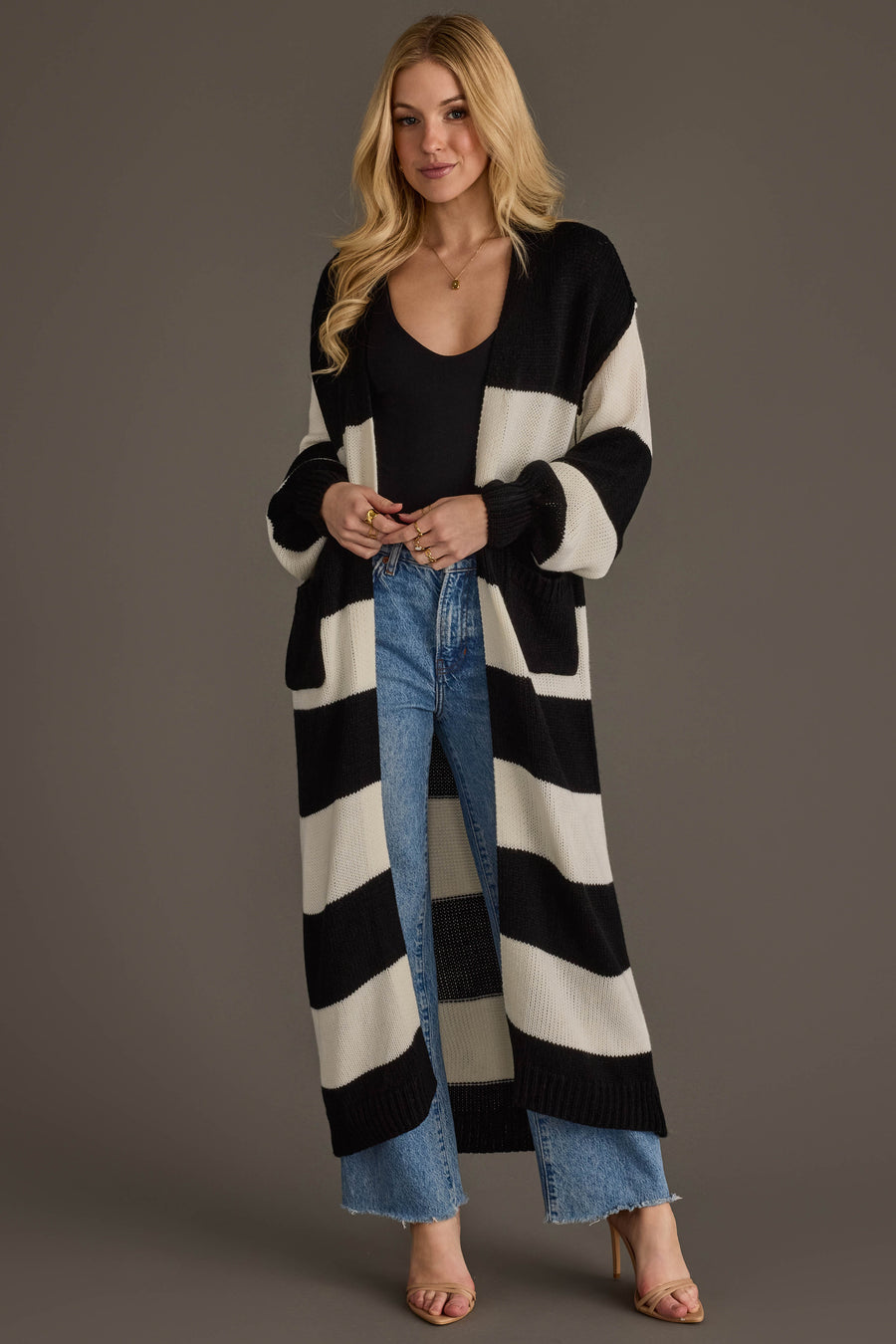 A woman models the Panache Apparel Collection Black & White Striped Duster Cardigan, layering it over a black top and blue jeans as she stands against a plain background with her hands clasped and a slight smile on her face.