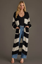A woman models the Panache Apparel Collection Black & White Striped Duster Cardigan, layered over a black top and blue jeans with sandals. She stands facing forward with her hands in her pockets against a plain gray background.