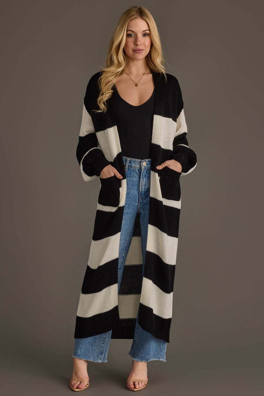 A woman models the Panache Apparel Collection Black & White Striped Duster Cardigan, layered over a black top and blue jeans with sandals. She stands facing forward with her hands in her pockets against a plain gray background.