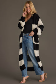 A woman smiles and poses against a plain gray background wearing the Panache Apparel Collection Black & White Striped Duster Cardigan over a black bodysuit, blue jeans, and tan heels.