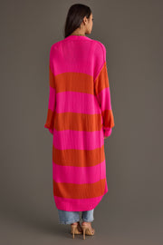 A woman with long brown hair, standing with her back to the camera against a plain background, is wearing the Panache Apparel Collection Bright Pink & Orange Striped Duster Cardigan, paired with blue jeans and beige heels.