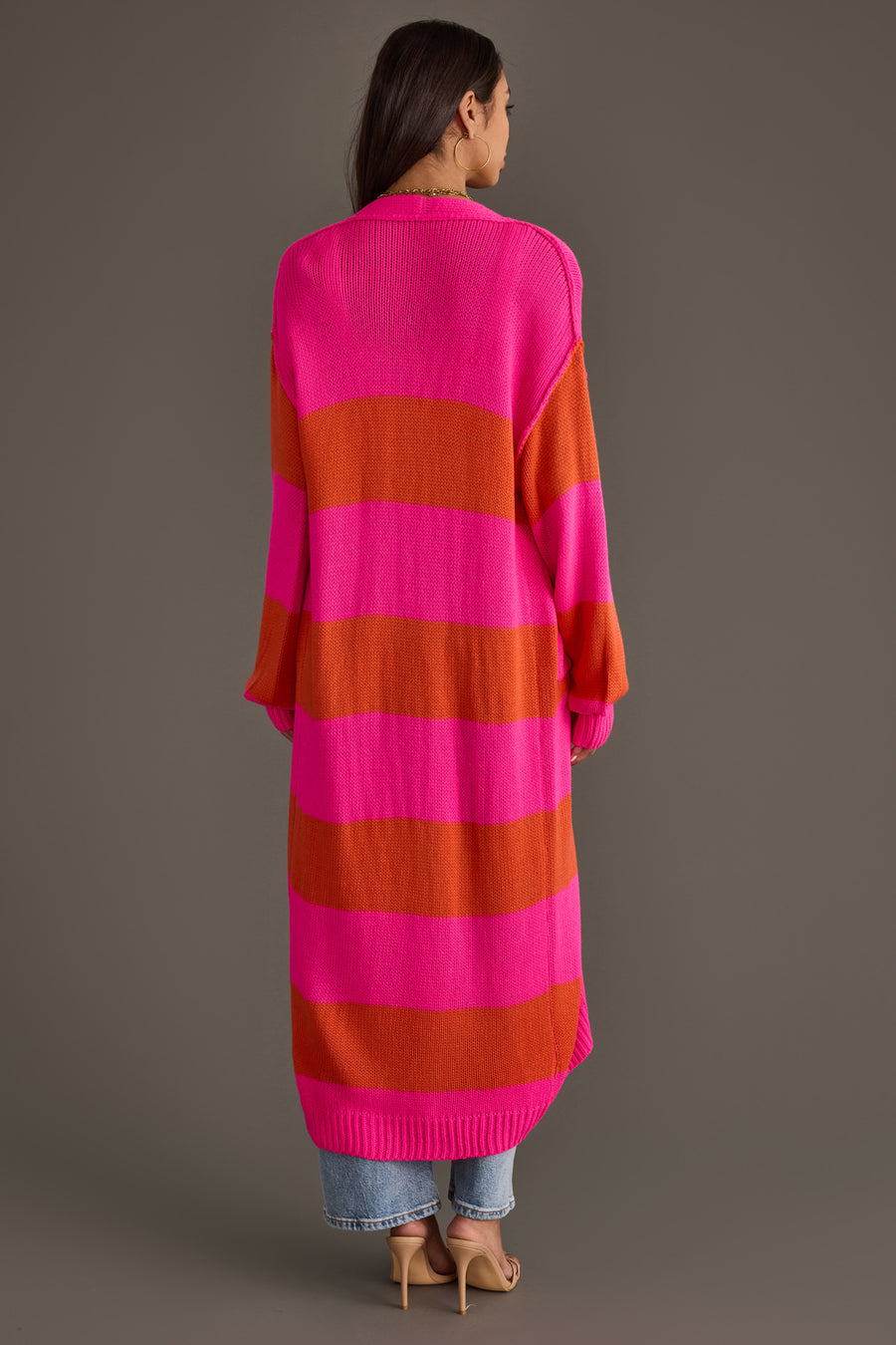 A woman with long brown hair, standing with her back to the camera against a plain background, is wearing the Panache Apparel Collection Bright Pink & Orange Striped Duster Cardigan, paired with blue jeans and beige heels.