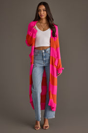 A woman stands wearing the Bright Pink & Orange Striped Duster Cardigan from Panache Apparel Collection, paired with a white ribbed crop top, light blue high-waisted jeans, and heeled sandals. She has long dark hair and layered necklaces, posing against a gray background.