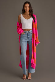 A woman stands smiling against a plain gray background, wearing a white tank top, light blue jeans, open-toed heels, and the Bright Pink & Orange Striped Duster Cardigan from Panache Apparel Collection.