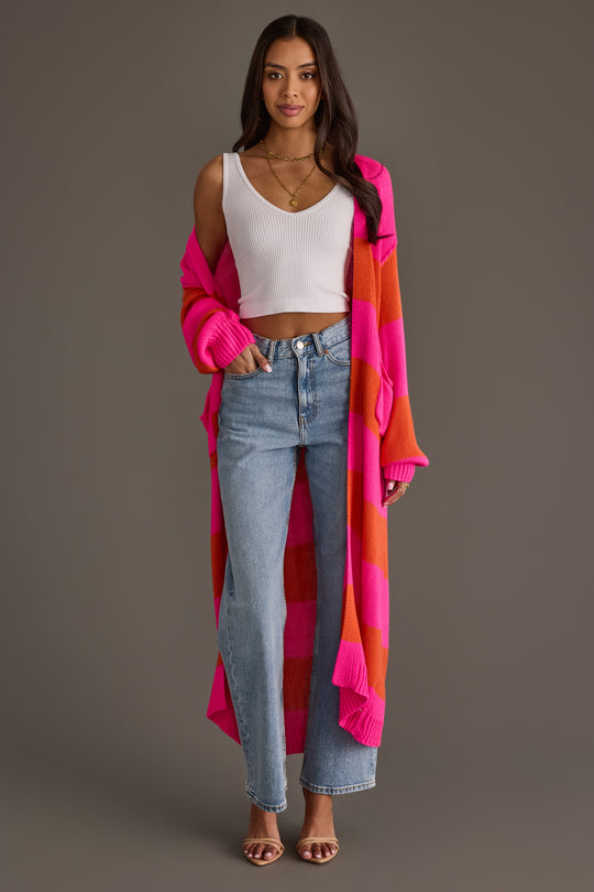 A woman stands smiling against a plain gray background, wearing a white tank top, light blue jeans, open-toed heels, and the Bright Pink & Orange Striped Duster Cardigan from Panache Apparel Collection.
