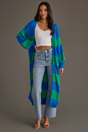 A woman stands against a plain background with one hand on her hip, wearing the Panache Apparel Collection Royal Blue & Green Striped Duster Cardigan layered over a white crop top, light blue high-waisted jeans, and heeled sandals.